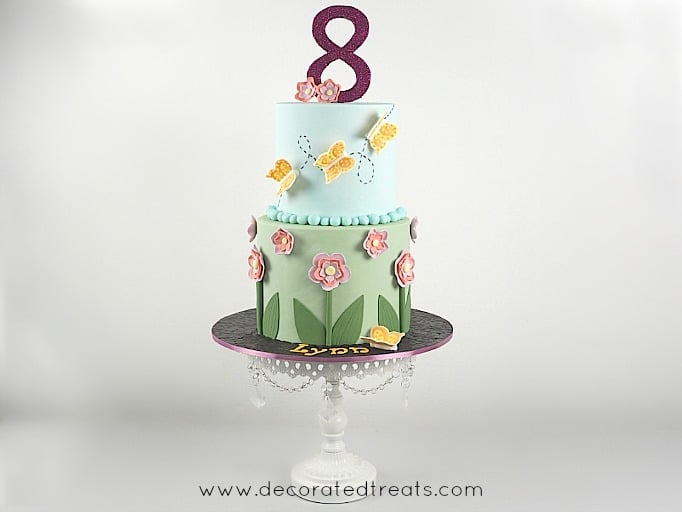 A 2 tier round cake in green and blue fondant. Bottom tier is decorated with fondant flowers and the top tier with royal icing butterflies in orange. Cake topper is a large glittery number 8