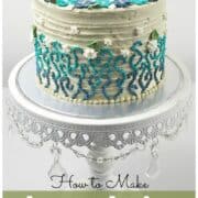 A round beige cake decorated with blue and turquoise buttercream piping and white flowers