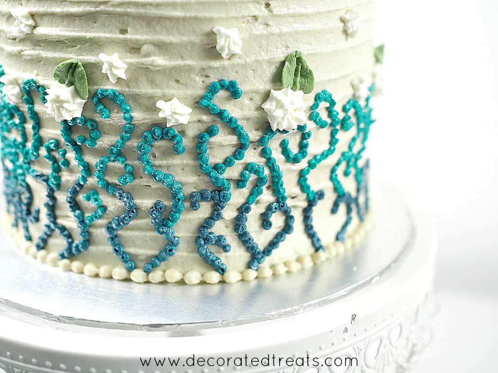 A round beige cake decorated with blue and turquoise buttercream piping and white flowers