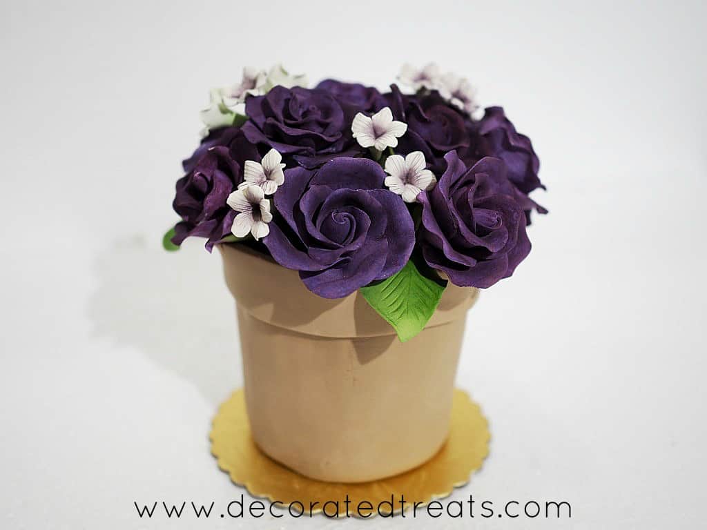 A flower pot shaped cake with purple roses and white filler gum paste flowers