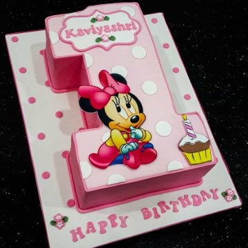 A number one shaped cake decorated with a Minnie Mouse baby image and a cup image. The cake is in pink with white polka dots while the cake board is white in pink polka dots
