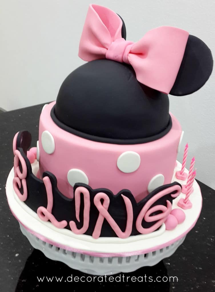A round pink cake in white polka dots with a Minnie head in black decorated with pink bow