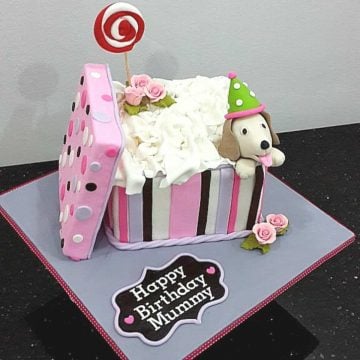 A square cake decorated like a gift box with its lid on the side a puppy topper at the corner of the box.