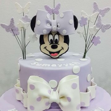 A single tier round purple cake with white polka dots, a Minnie Mouse face topper and butterflies and flowers side toppers. The cake has a large white bow on the front.
