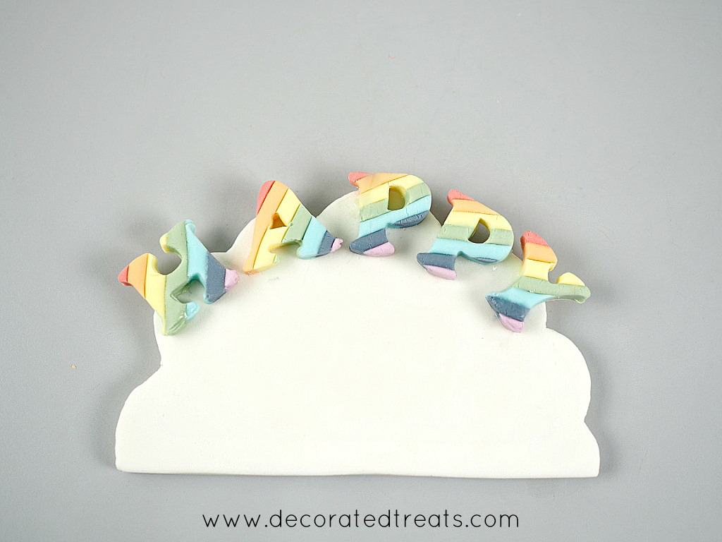 The word HAPPY in rainbow colors on a piece of cloud shaped white fondant.