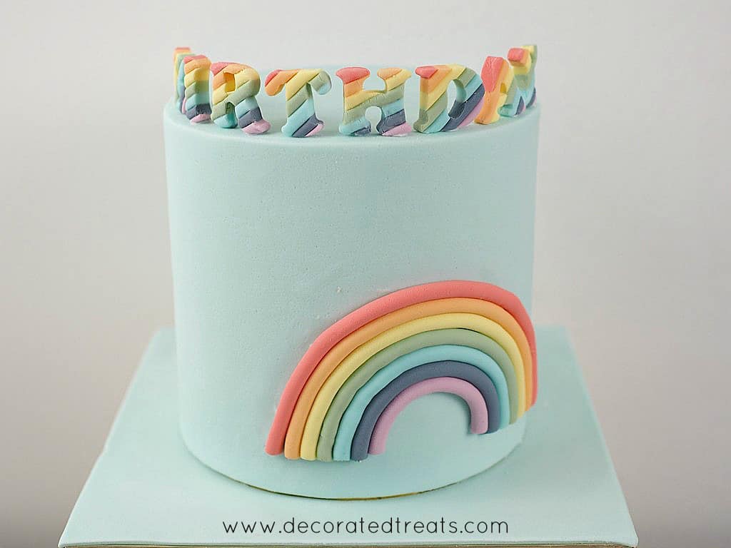 A blue cake decorated with 2D rainbow on the sides and rainbow alphabets on top