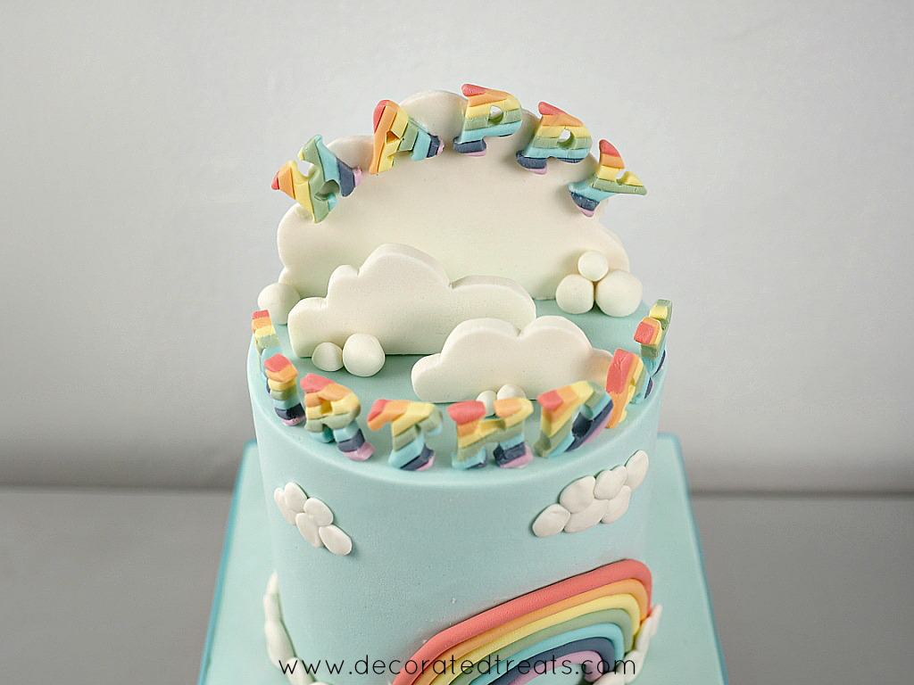 A round blue cake decorated with rainbow and clouds on the sides and rainbow alphabets on top of the cake