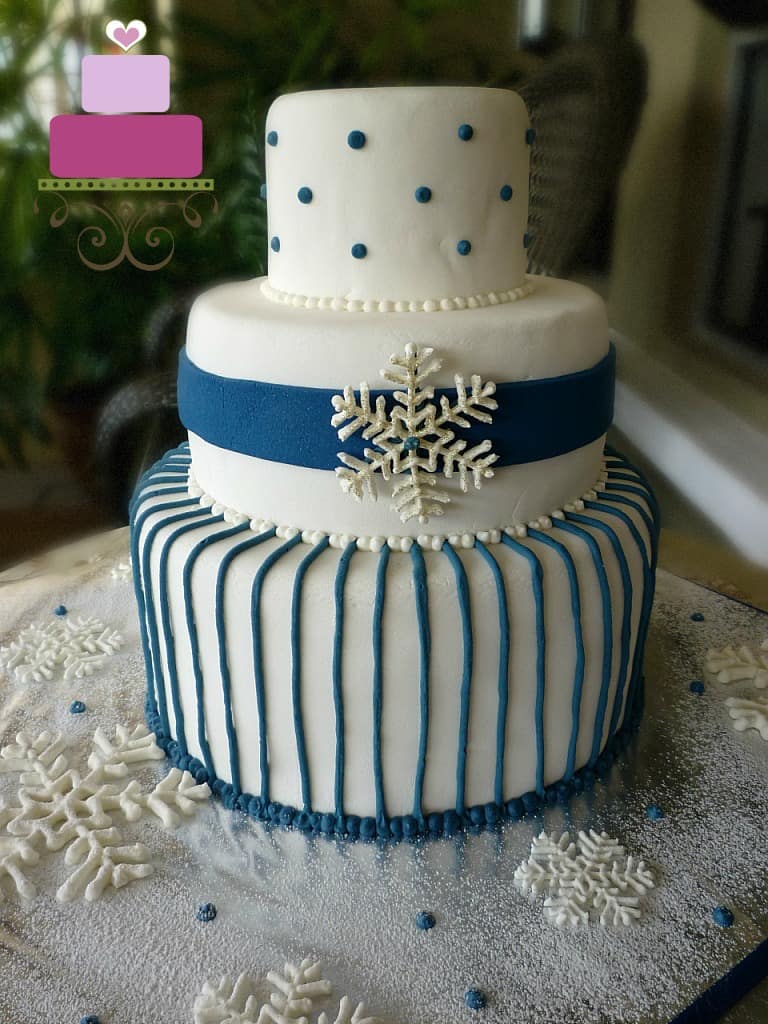 A three tier white cake decorated with blue stripes and white royal icing snowflake.