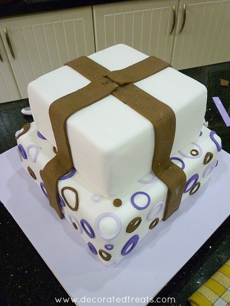 A two tier cake with circle purple and brown cut outs on the bottom tier and fondant strips on the top tier