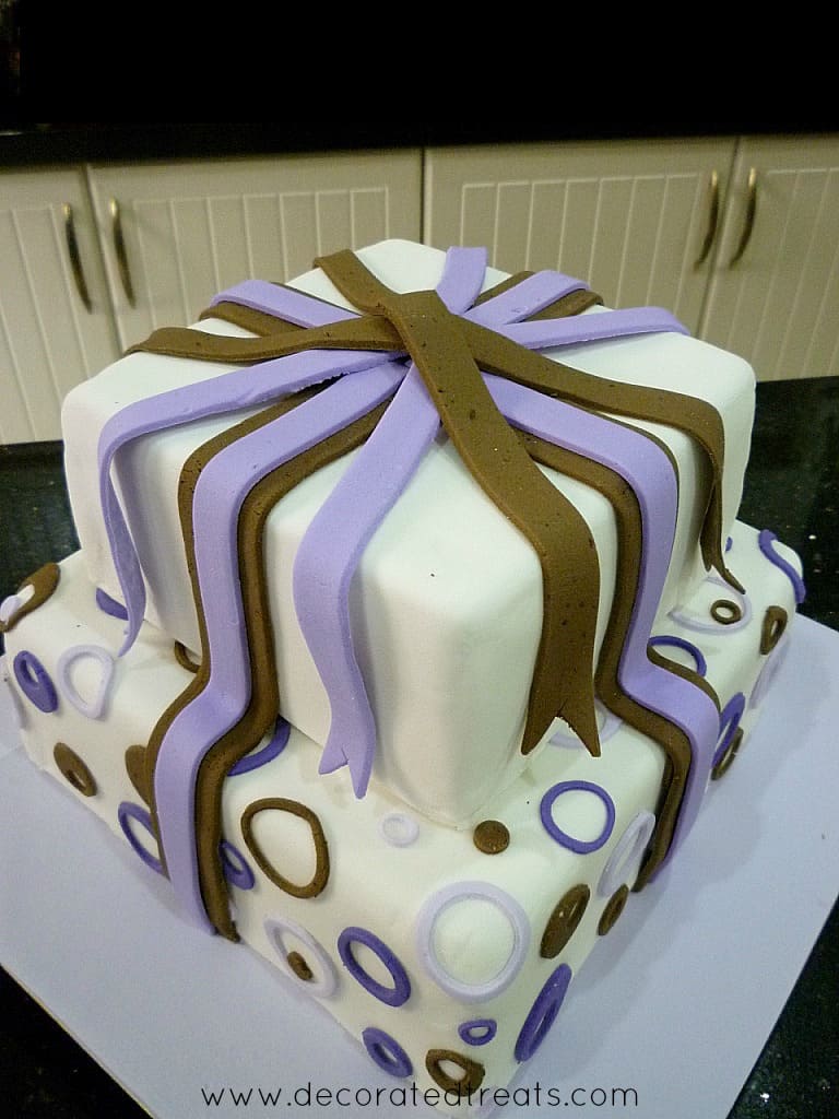 A two tier cake with circle purple and brown cut outs on the bottom tier and fondant strips on the top tier