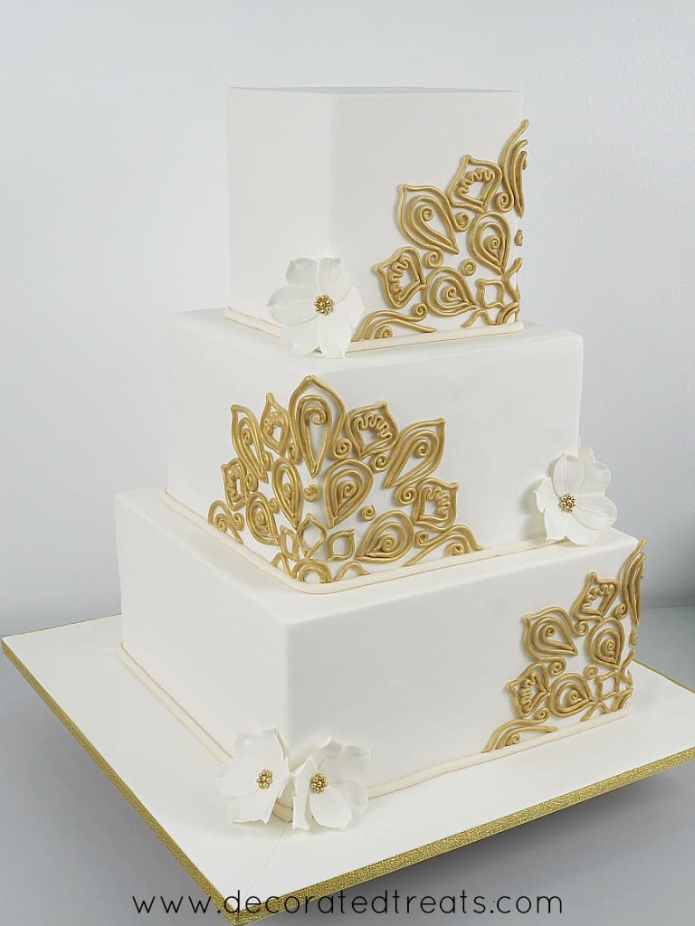 A 3 tier square cake in white fondant and gold lace and white gum paste flowers
