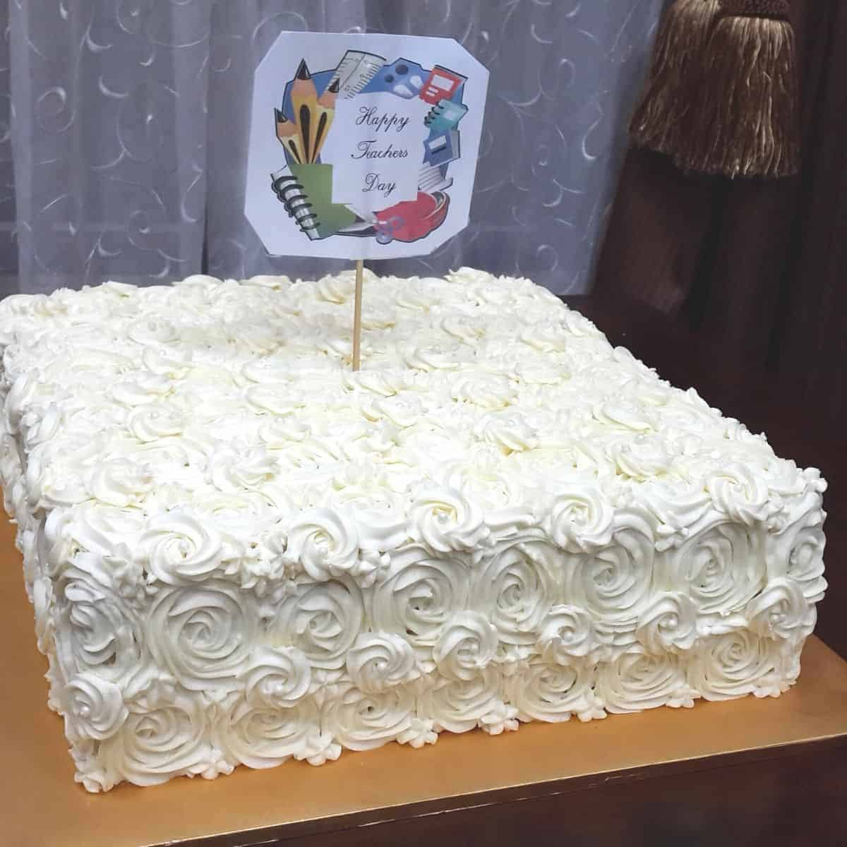 A square cake decorated in white buttercream rosettes and a Teachers Day cake topper
