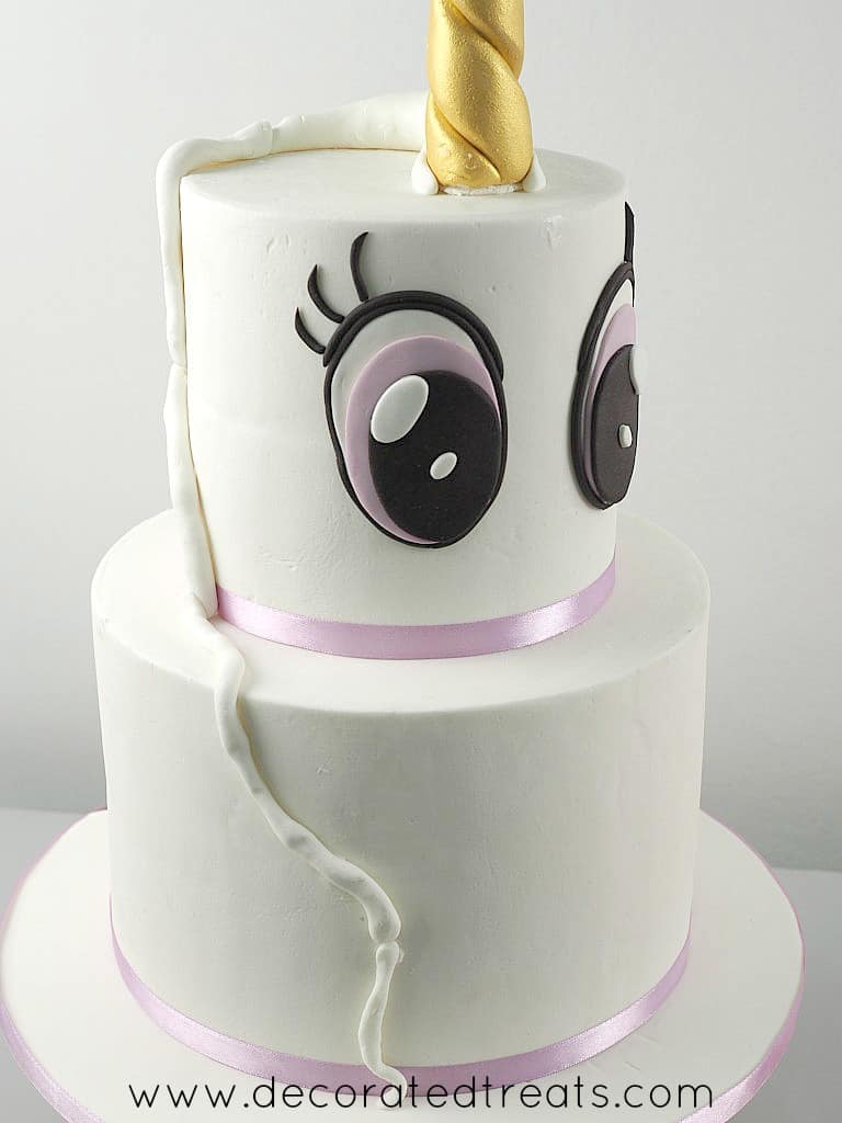 A two tier cake with gold unicorn horn topper and large fondant eyes on the top tier. A long strip of fondant is attached to the cake