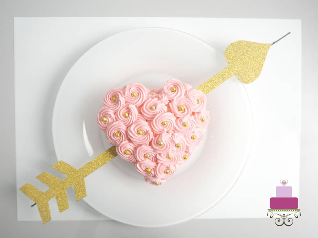 A mini heart shaped cake covered in pink buttercream rosettes and gold beads and a paper heart going through the sides