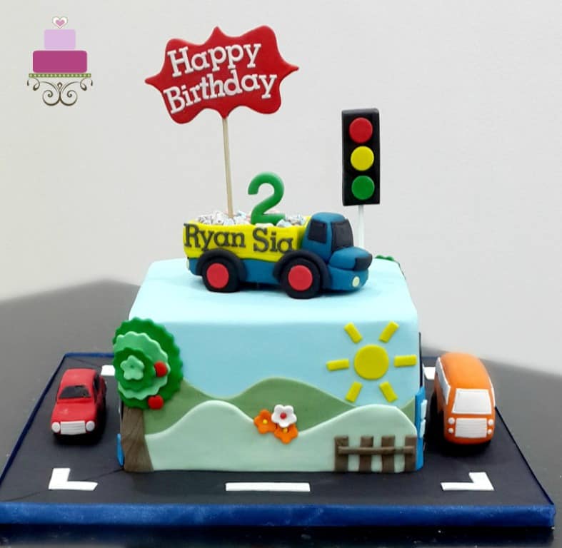 A square cake with a dump truck cake topper. Cake is decorated with scenery on the sides and a fondant car and bus on the cake board.