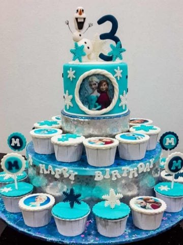 A blue round cake with 3d Olaf topper on a cupcake stand, with cupcakes on the stand