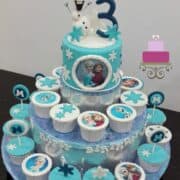 A blue round cake with 3d Olaf topper on a cupcake stand, with cupcakes on the stand