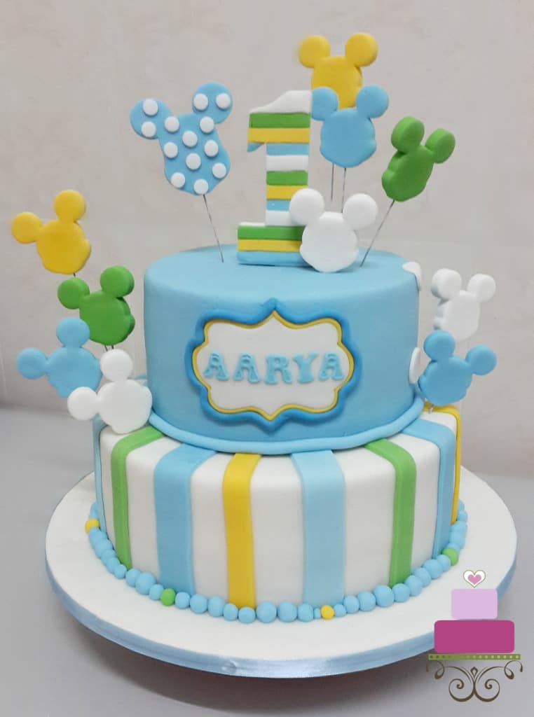 A 2 tier cake decorated with Mickey face cut outs in blue, white, green, yellow and white and a matching number 1 topper