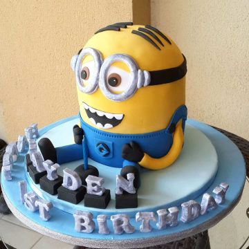 A Minion shaped birthday cake with silver alphabets on a blue cake board.