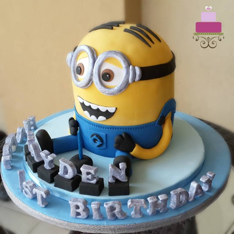 A Minion shaped birthday cake with silver alphabets on a blue cake board