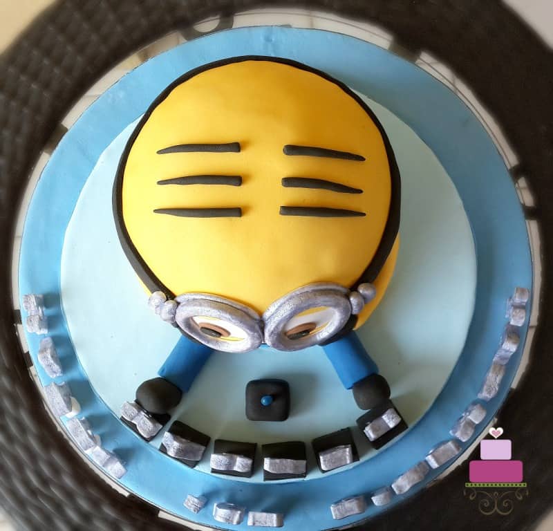 A Minion shaped birthday cake with silver alphabets on a blue cake board.