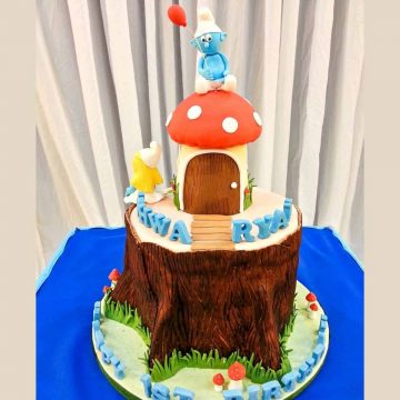 A mushroom on a tree stump cake decorated in Smurfs theme. A 3D Smurf in fondant is the cake topper.