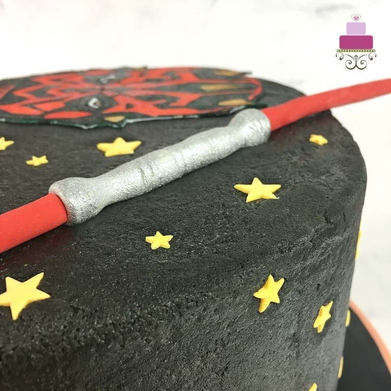 A round cake decorated with Darth Maul face image and lightsaber topper