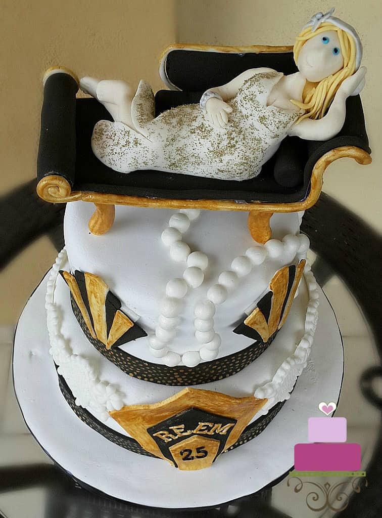 A two tier cake in black, white and gold with a lady figurine lying down on a couch.