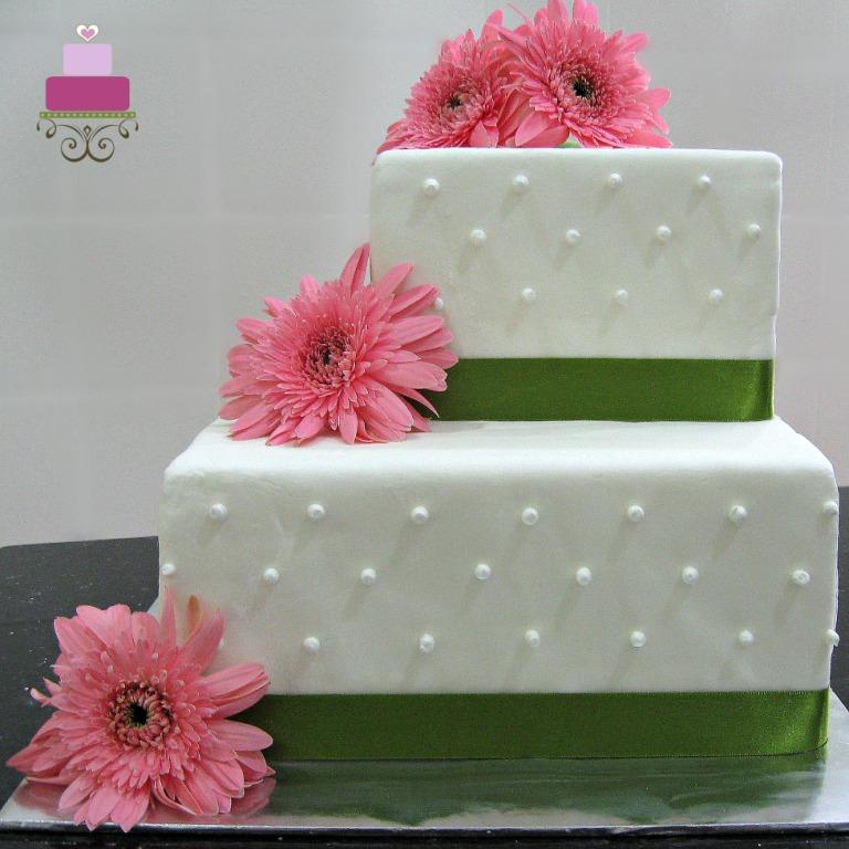 A 2 tier square wedding cake decorated with green ribbon border and fresh pink gerbera flowers