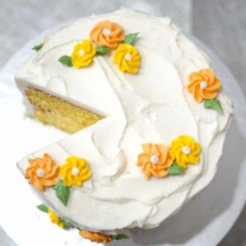 A round cake decorated with white buttercream and orange and yellow piped buttercream flowers. A slice of the cake is cut out