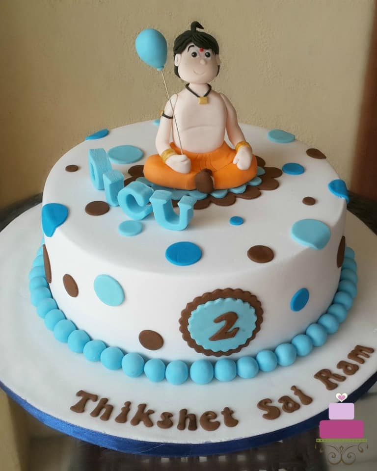 A round cake with blue and brown polka dots and Chhota Bheem cake topper holding a blue balloon.