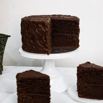 A chocolate cake on a white stand with a portion cut out.