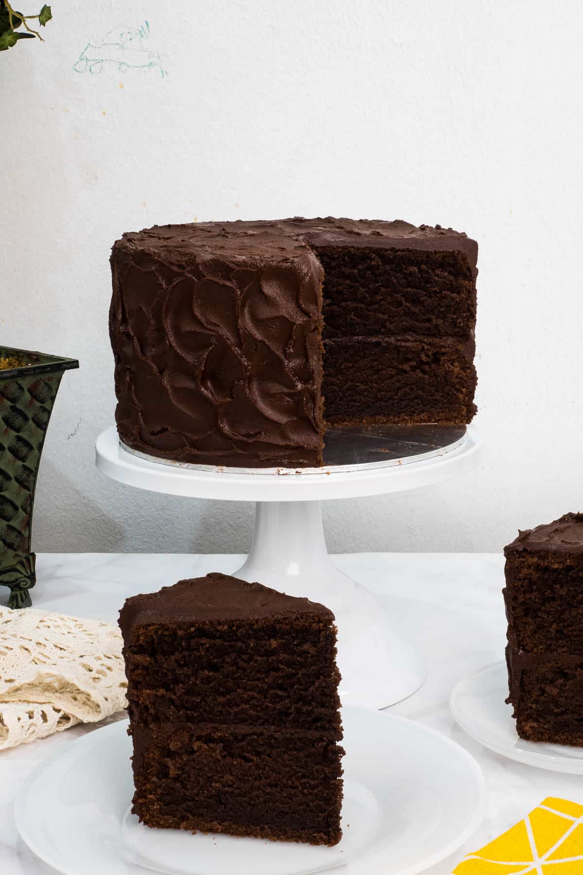 A chocolate cake on a white stand with a portion cut out and 2 slices of the cake on white plates on the front and side.