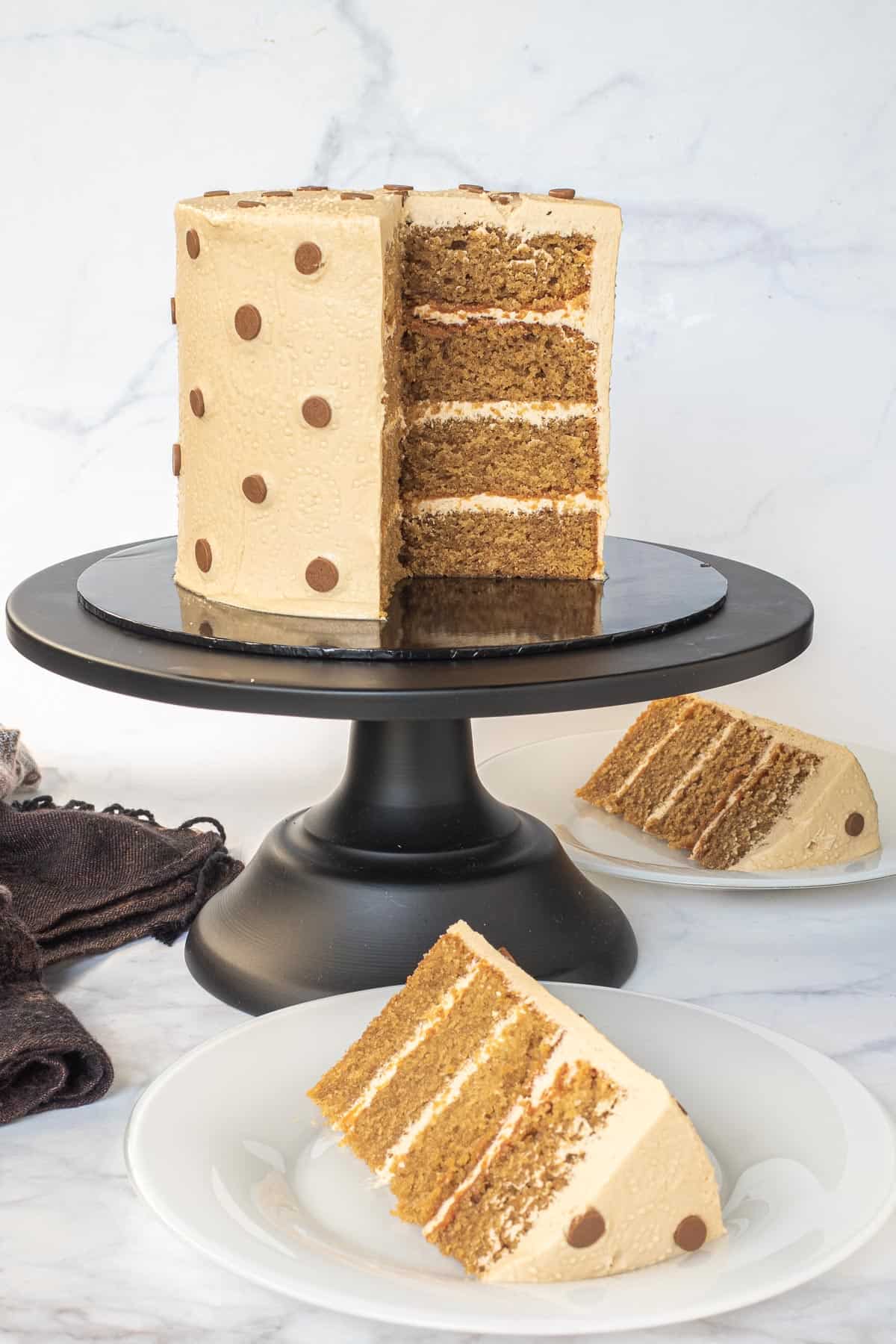 A round coffee flavored cake decorated with polka dots icing. Cake is in a black cake stand with a slice cut onto a white plate.