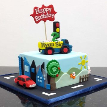 A square cake with a dump truck cake topper. Cake is decorated with scenery on the sides and a fondant car on the cake board.
