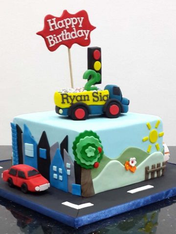 A square cake with a dump truck cake topper. Cake is decorated with scenery on the sides and a fondant car on the cake board.