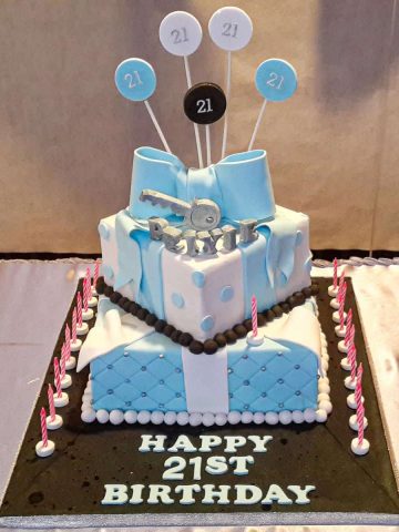 A 2 tier square birthday cake with a large blue bow and silver key topper.