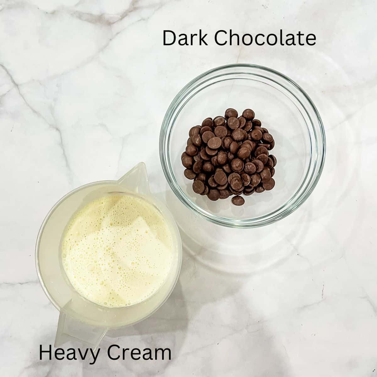 A bowl of dark chocolate chips and jug of cream against marble background.