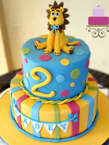 A two tier cake in yellow and blue with a lion cake topper and a large number 2 on the top tier.