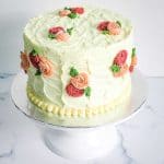 A round cake covered in white buttercream with pink and maroon rosettes and green leaves. Cake is on a white cake stand.
