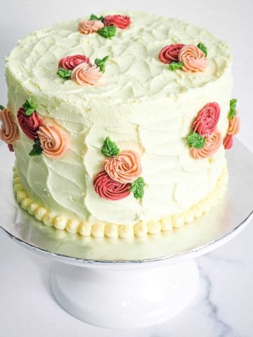 A round cake covered in white buttercream with pink and maroon rosettes and green leaves. Cake is on a white cake stand