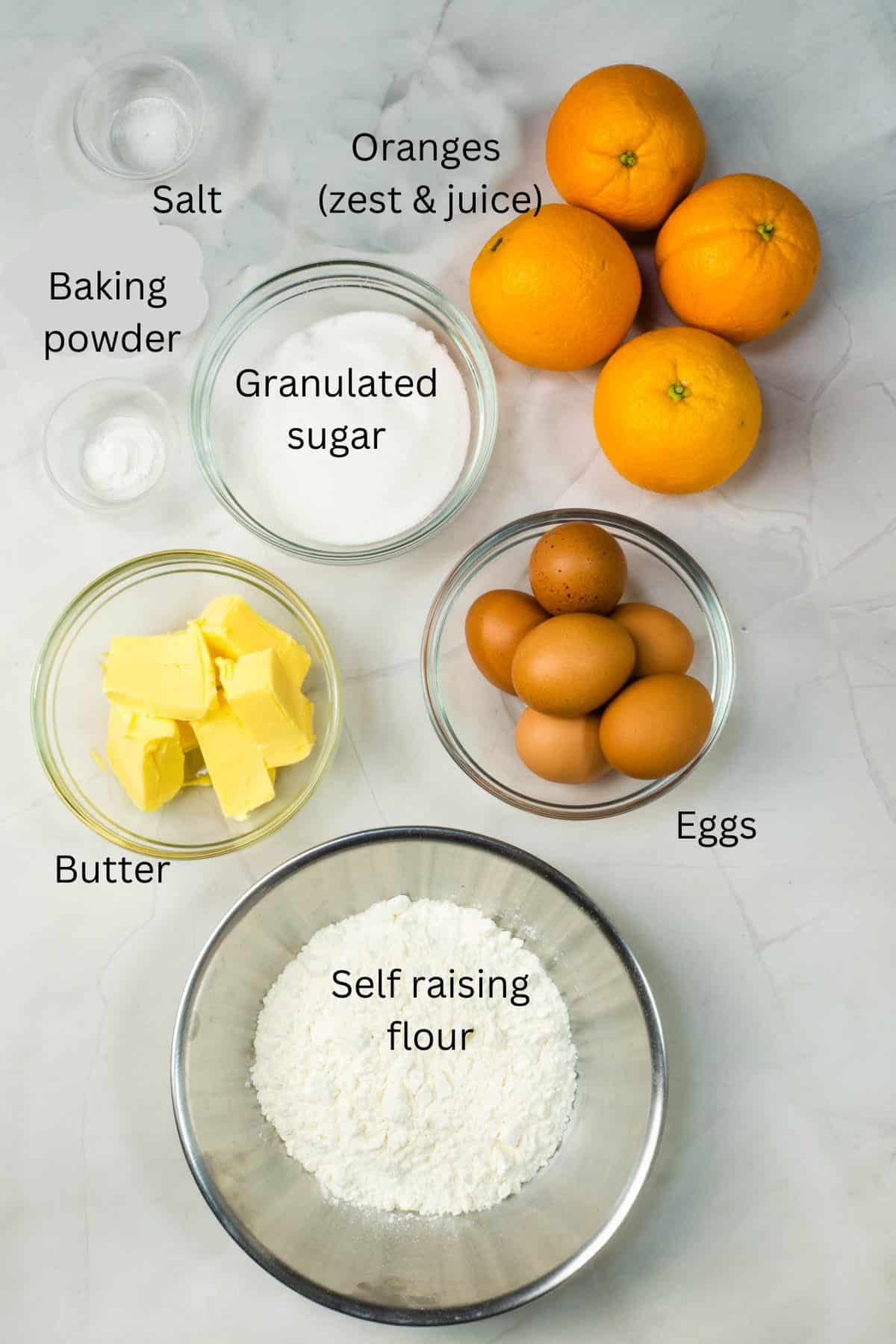 Self raising flour, granulated sugar, butter, eggs, oranges, baking powder and salt in bowls against a marble background.