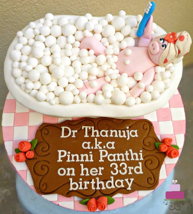 A bath tub shaped cake with a 3D fondant pig holding a brush in it. Cake is topped with fondant bubbles and placed on a pink and white checked cake board with a large brown birthday wish plaque