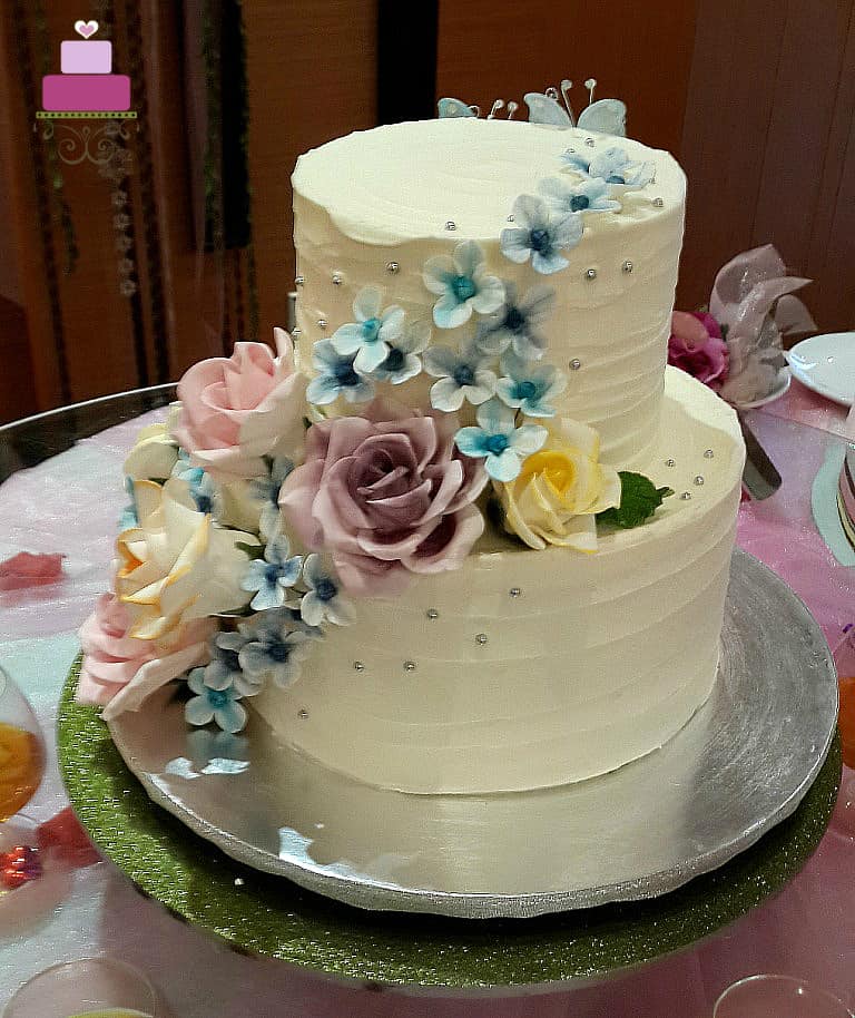 A two tier wedding cake with rustic buttercream deco, pink, purple and yellow roses and blue hydrangeas