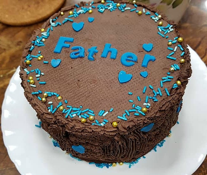 A round cake covered in piped chocolate icing, with blue alphabets spelling 'Father'. Cake is decorated with blue hearts and sprinkles in blue and gold.