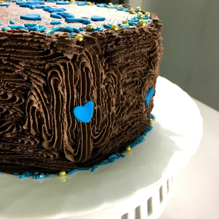 A round chocolate cake covered in chocolate icing and decorated with blue hearts and sprinkles.