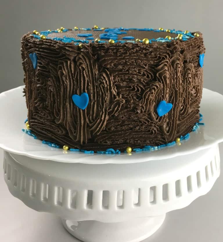 A round chocolate cake covered in chocolate icing and decorated with blue hearts and sprinkles.