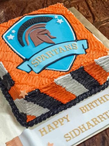 A square cake with orange, black and white side strips and a Spartans logo.
