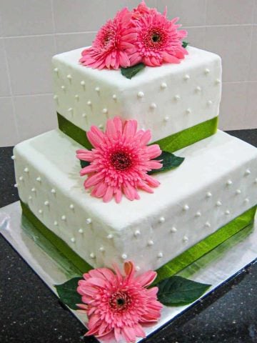 A 2 tier square wedding cake decorated with green ribbon border and fresh pink gerbera flowers