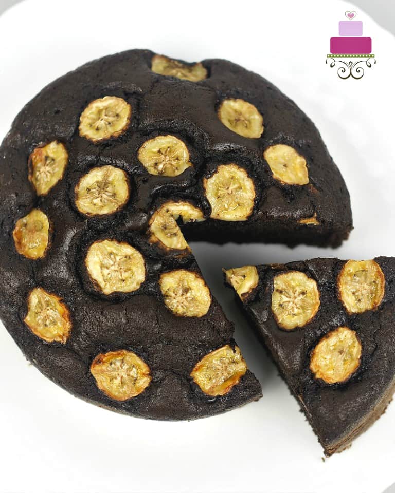 A round banana and chocolate cake topped with caramelized banana slices. A slice of the cake is cut out.